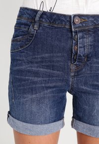 Tummansiniset farkkushortsit, joissa on käännetyt lahkeet, etutaskut, kontrastiväriset tikkaukset sekä nappisuljenta pronssinvärisillä napeilla.