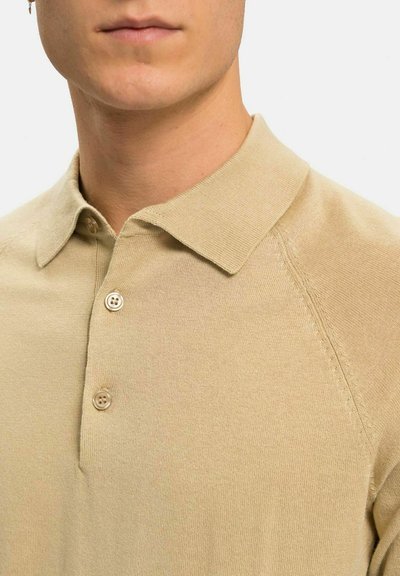 Beige pikétröja med mjuk textur, som har en krage, två knappar och en figurnära design. Inkluderar subtila detaljer med raglanärmar.