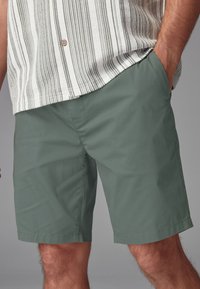 Pantaloni corti verdi realizzati in un tessuto morbido e leggero con una texture liscia. Presentano tasche laterali e un orlo dritto.