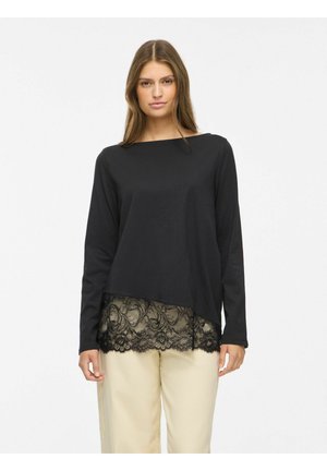 Femme portant un haut noir à manches longues avec un bord en dentelle à l'ourlet, assorti à un pantalon beige clair sur un fond uni.
