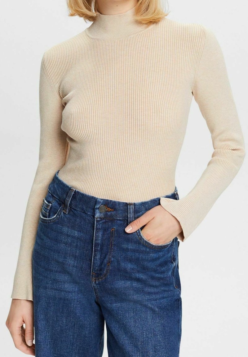 Béžový žebrovaný rolák s dlouhým rukávem, spárovaný s tmavě modrými džínami s knoflíkovým zapínáním a bočními kapsami.