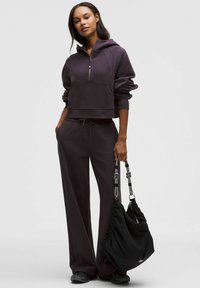 Lila kurzes Hoodie mit halbem Reißverschluss und Vordertasche, kombiniert mit passenden weiten Hosen. Schwarze Schultertasche mit grauen Griffen.