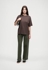 Hnědé bavlněné tričko s volným střihem, spárované se zelenými širokými kalhotami. Modelka drží malou černou kabelku a nosí černé platformové boty.