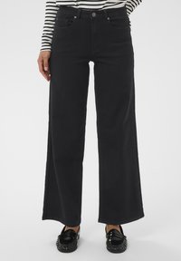 Personne portant un pantalon large noir, des mocassins noirs et un haut à rayures noir et blanc à manches longues, se tenant avec une main près de la poche.