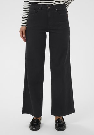 Osoba w czarnych szerokich dżinsach, czarnych loafersach i długim rękawie w biało-czarną paski, stoi z jedną ręką blisko kieszeni.