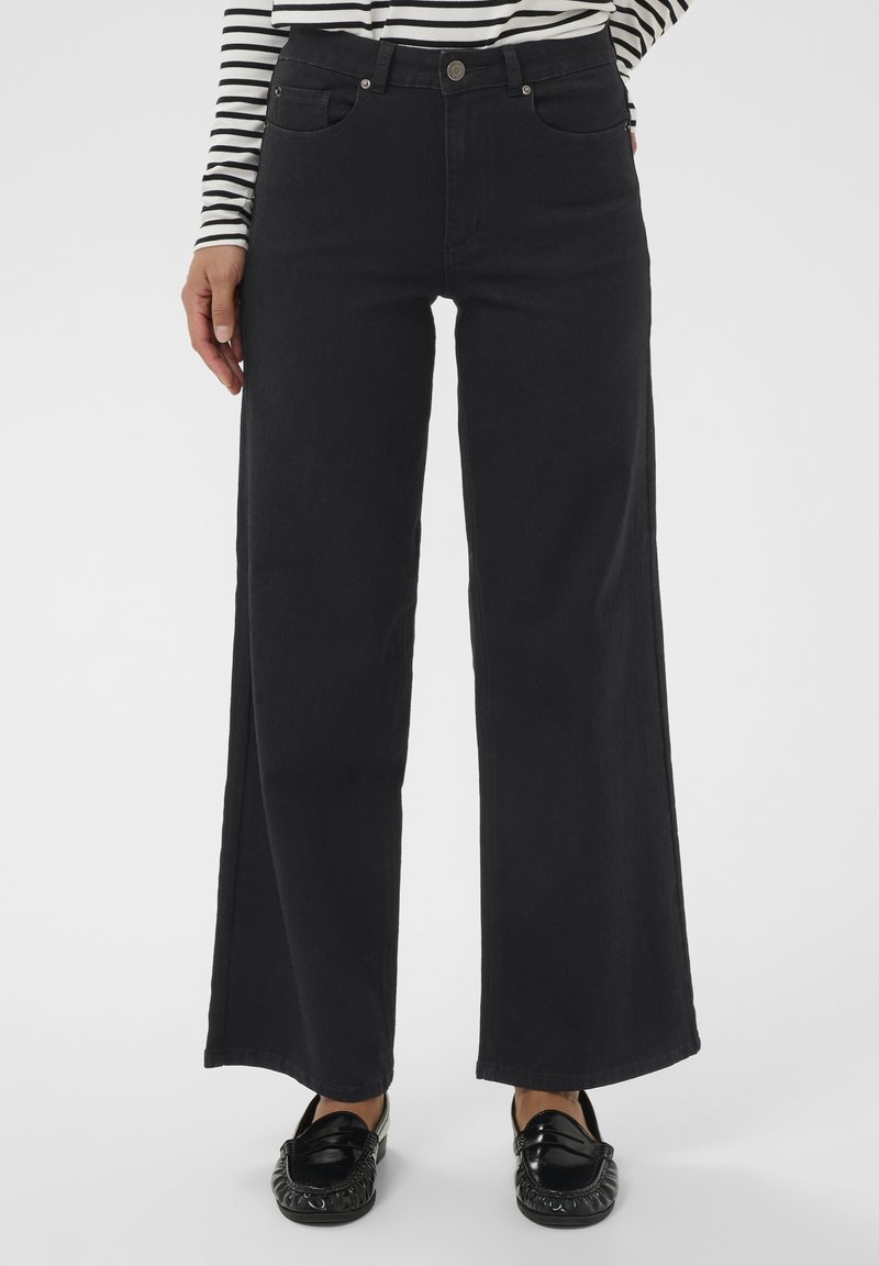 Personne portant un pantalon large noir, des mocassins noirs et un haut à rayures noir et blanc à manches longues, se tenant avec une main près de la poche.