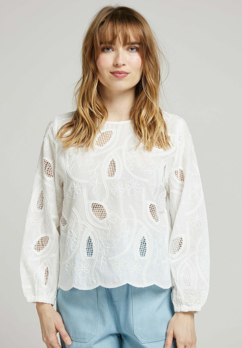 Blouse blanche brodée avec de longues manches ballon, un ourlet festonné, des motifs floraux et de feuilles, présentant des découpes transparentes sur l'ensemble.