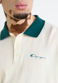 Kremfarget ribbestrikket polo skjorte med grønn krage, utstyrt med tre knapper og en blå brodert "Champion"-logo på brystet.