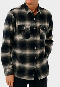 Camisa de franela a cuadros en blanco y negro con dos bolsillos en el pecho, cuello abotonado y mangas largas con un ajuste holgado y tejido texturizado.