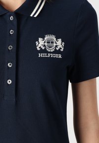 Tamno plava polo majica s bijelim izvezenim logotipom i gumbima. Rebrasti ovratnik s dvije bijele pruge, koji nudi izgled teksturirane tkanine.