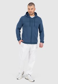 Felpa blu con zip, pannello superiore testurizzato, cappuccio con cordoncino e tasche frontali, abbinata a pantaloni sportivi bianchi e sneakers grigie.
