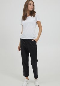 Hvid kortærmet t-shirt med rund halsudskæring, kombineret med sorte tapered bukser i faux læder og hvide sneakers, stående med den ene hånd i lommen.