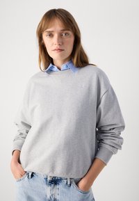 Felpa grigia con polsini e orlo a coste, con logo ricamato. Indossata sopra una camicia colletto azzurra. Jeans in denim chiaro.