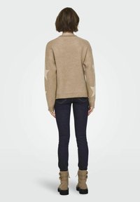 Beige gebreide sweater met sterrenpatronen op de mouwen, gecombineerd met donkere jeans en tan enkellaarsjes. Geweven stof, ontspannen ontwerp, ribgebreide manchetten.