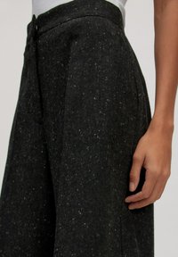 Schwarze, strukturierte Hose aus Wollmischung, mit hoher Taille, Falten und entspannter Passform. Seitentasche sichtbar an der Hüfte.