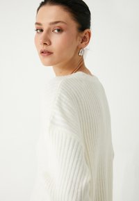 Biały sweter z ribbowanym splotem, długimi rękawami, z szerokim dekoltem i miękką fakturą. Podkreślony srebrną kolczykiem w uchu modelki.