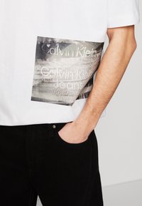 Bílé bavlněné tričko s černou a šedou potiskovou nášivkou zobrazující "Calvin Klein Jeans" v texturovaných písmenech. Černé kalhoty s přední kapsou.