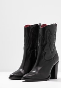 Une paire de bottines en cuir noir avec des talons épais, des bouts pointus, des fermetures éclair latérales et des motifs de foudre cousus.