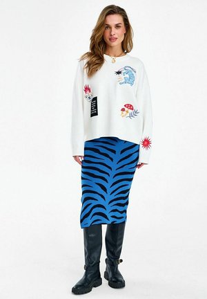 Kobieta w białym swetrze z kolorowymi naszywkami, spódnicy w niebiesko-czarne paski zebry i czarnych wysokich botkach do kolan, stojąca na tle białego tła.