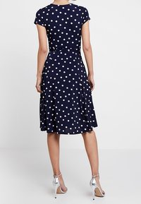 Granatowa sukienka w groszki z krótkimi rękawami, dopasowaną talią, rozkloszowaną spódnicą i subtelną fakturą. Do tego srebrne sandały na wysokim obcasie.