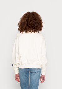 Persona con cabello rizado castaño, vistiendo un sudadera blanca y jeans azules, de pie frente a un fondo claro y liso.