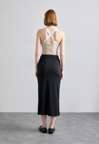 Beige top zonder mouwen met gekruiste witte banden en een lage rug, gecombineerd met een strakke zwarte midi-rok met zijsplitten en een gladde textuur.