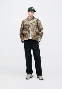 Μπουφάν με καμουφλάζ μοτίβο σε καφέ και πράσινο, συνδυασμένο με γκρι πουκάμισο και μαύρο παντελόνι. Διαθέτει τέσσερις μπροστινές τσέπες. Γκρι αθλητικά παπούτσια ολοκληρώνουν την εμφάνιση.