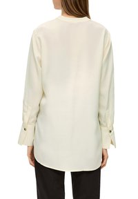 Langärmelige, cremefarbene Bluse mit glatter Textur, die über geknöpfte Manschetten und einen abgerundeten Saum verfügt, von hinten gezeigt.