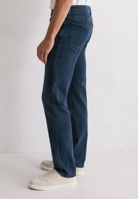 Personne portant un jean droit bleu marine et des baskets blanc cassé sur un fond clair uni, vue de côté.