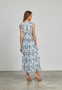 Abito maxi floreale bianco e blu, senza maniche, con busto aderente e gonna fluente, in tessuto leggero con un design plissè, abbinato a tacchi beige.