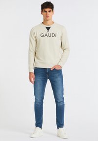 Felpa beige chiaro con testo nero "GAUDÌ" e design a triangolo; abbinata a jeans blu e sneakers bianche su un modello.