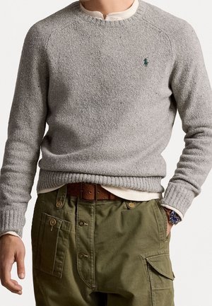 Man draagt lichtgrijze gebreide trui met donkergroene geborduurde logo, wit ondershirt, olijfgroene knoopbroek, bruine riem en polshorloge.