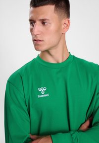 T-shirt verde a maniche lunghe in tessuto morbido con scollatura rotonda e logo ricamato bianco sul petto. Design semplice e dritto.