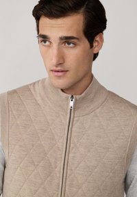 Bež prsluk s kvadratnim uzorkom, visokim ovratnikom i srebrnim zatvaranjem, s pletenim rebrastim naglasima i glatkom sivom tkaninom ispod.