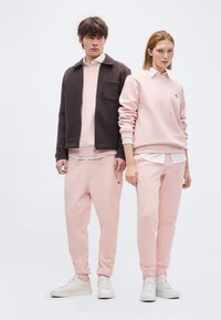 Νεαρός άνδρας και γυναίκα στέκονται, φορώντας συντονισμένο ανοιχτό ροζ φόρμες και φούτερ, λευκά αθλητικά παπούτσια και λευκά πουκάμισα με γιακά.