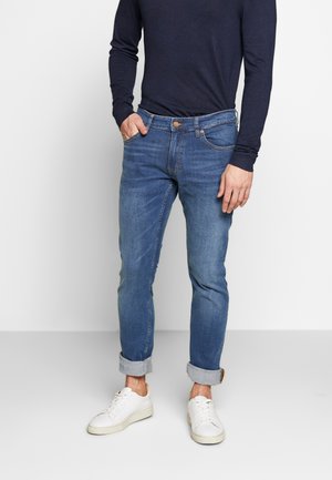 Mann trägt hochgekrempelte blaue Jeans, weiße Sneaker und ein marineblaues Langarmshirt, steht mit einer Hand in der Tasche.