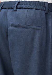 Κοντινό πλάνο σκούρων μπλε παντελονιών με ελαστική μέση, θηλιές για ζώνη και μια πίσω τσέπη με καδράρισμα.