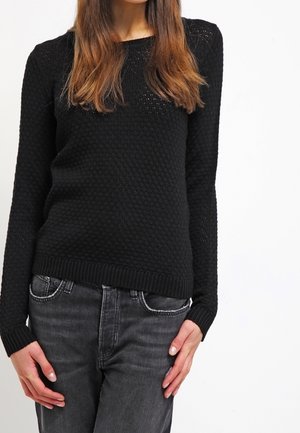 Vrouw in een zwarte, gestructureerde gebreide trui en donkergrijze jeans, staand met handen bij haar heupen tegen een effen witte achtergrond.