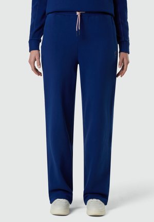 Persona con pantalones anchos azul marino con un cordón rosa claro y zapatillas blancas, de pie contra un fondo liso.