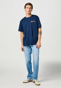 Mörkblå t-shirt med kort ärm med texten "WRANGLER", tillsammans med ljusblå jeans och vita skor med svarta ränder.
