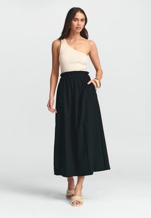 Zwarte maxi-jurk met een gathered elastische taille, gecombineerd met een beige ribbelige één-schoudertop. Model draagt gevlochten sandalen, minimalistische accessoires.
