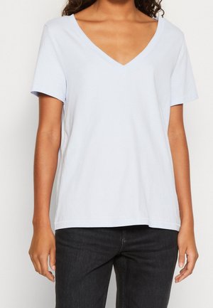 Kobieta ubrana w jasnoniebieską koszulkę z krótkim rękawem i dekoltem w serek oraz ciemne dżinsy, stojąca na tle jednolitego białego tła.