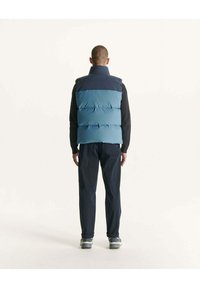 Blauwe puffervest met een lichterblauwe bovenkant, gecombineerd met donkere broeken. Zachte textuur, horizontale secties en hoge kraag zijn zichtbaar.