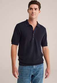 Vīrietis, kas ģērbies tumšā tekstūrā īsās piedurkņu polo kreklā un zilās džinsās, stāvot pretī vienkāršai gaišai fona un skatoties pa labi.