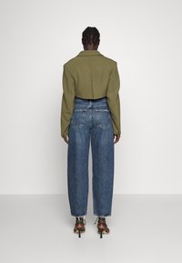 Giacca corta verde oliva con spalline, abbinata a jeans in denim azzurro a vita alta con gamba larga e sandali con tacco marroni con dettagli in catena.