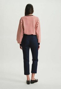 Chemise à rayures rouges et blanches à manches longues avec des poignets noirs, associée à un pantalon tailleur bleu marine et des chaussures noires, vue de dos.