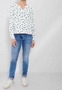 Person iført en hvid langærmet bluse med mørke paisley-mønstre, blå optrækkede jeans og hvide sneakers, stående mod en lys grå baggrund.