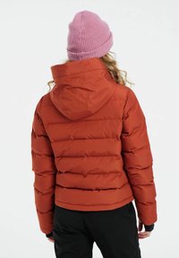Rød pufferjakke med syede, vandrette sektioner, hætte og elastiske manchetter. Bæres med en lys pink strikket hue. Sorte bukser er synlige.