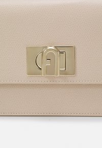 Borsa a mano in pelle beige con superficie testurizzata, dotata di una chiusura dorata con logo in rilievo. Le cuciture lungo i bordi ne migliorano la resistenza.