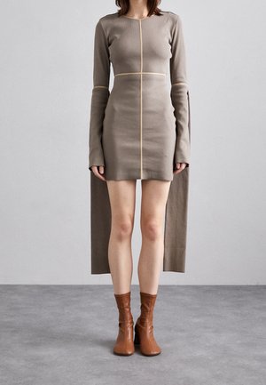 Vrouw draagt een aansluitende taupekleurige mini-jurk met lange verlengde mouwen en bruine enkellaarsjes, staand op een grijze vloer.